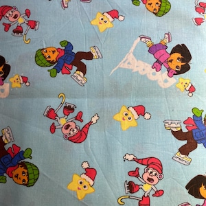 May include: Light blue fabric featuring cartoon characters ice skating. The design includes Dora the Explorer, a monkey, and star shapes wearing Santa hats. The characters are in winter attire, with a snowy background.