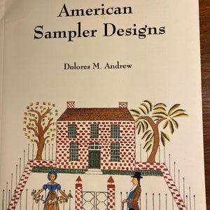 May include: A book cover with the title "American Sampler Designs" by Dolores M. Andrew. The cover features a colourful illustration of a brick house with a red roof, two trees, a fence, a gate, a woman holding flowers, a man with a cane, and a black dog.