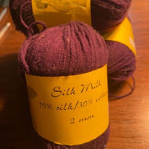 May include: Three skeins of deep purple yarn with a yellow label that reads "Silk Milk 70% silk/30% cotton 2 oz"