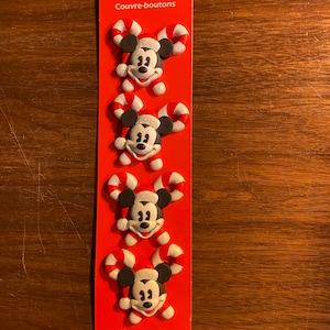 May include: A pack of four Christmas-themed Mickey Mouse button covers. The covers are red, white and black and feature Mickey Mouse wearing a Santa hat and holding candy canes.