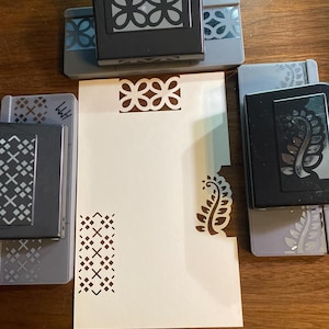 Puede incluir: Una colección de perforadoras de papel y una tarjeta crema con diseños decorativos recortados. Las perforadoras son negras y grises, con patrones florales y geométricos. La tarjeta muestra detalles recortados a juego, mostrando suministros de manualidades.