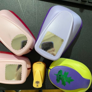 May include: A selection of craft punches in shades of pink, purple, and yellow. The punches have different shapes, including a scalloped circle, a scalloped square, and a leaf. A yellow punch features a palm tree design.