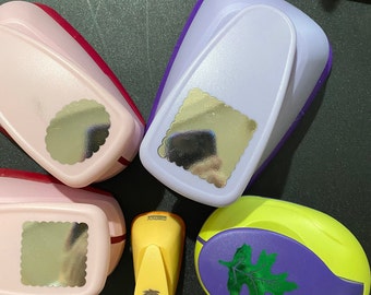 May include: An assortment of craft punches in various colors, including pink, purple, and yellow. The punches have different shapes, such as a scalloped circle, a scalloped square, and a leaf. A yellow punch features a palm tree design.