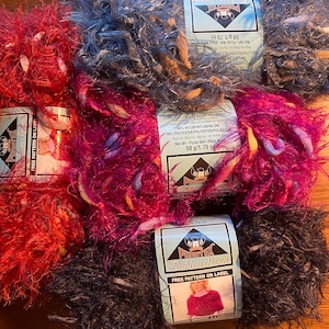 May include: A collection of colorful Phentex Fashion Seven yarn skeins. The yarn is available in shades of red, gray, pink, and black. Each skein is labeled with the brand name and a free pattern. Each skein weighs 1.75 oz.