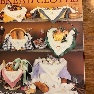 May include: A brochure titled "Holiday Bread Cloths" featuring nine designs. The cover displays various baskets with bread, each lined with decorative cloths. The cloths are embroidered with holiday-themed motifs, including a snowman, Santa, and a leprechaun.