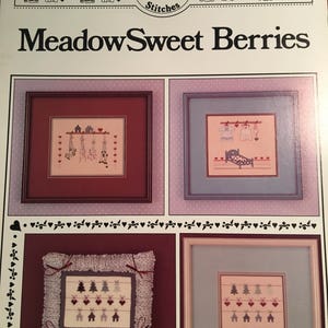 May include: A cross stitch pattern booklet titled "Meadow Sweet Berries" by Country Berry Stitches. The booklet features four cross stitch designs, each framed in a different color. The designs include a row of houses with hanging flowers, a row of houses with hanging clothes, a row of trees with hanging hearts, and a row of trees with hanging hearts.