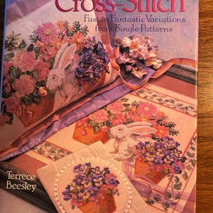 Op de afbeelding: Boekomslag voor "Progressive Cross-Stitch: Fast to Fantastic Variations from Single Patterns" door Terrece Beesley. De omslag toont verschillende kruissteekontwerpen met bloemen- en konijnenmotieven.