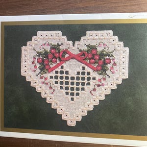 May include: A heart-shaped cross-stitch design with a lattice center, adorned with red berries, green leaves, and a red bow. The design is set against a dark green background with a gold border, creating a vintage aesthetic.
