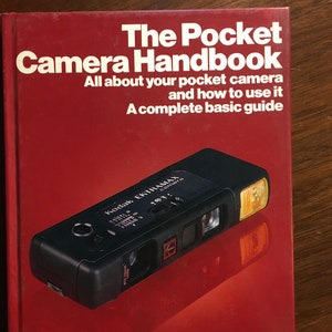 Puede incluir: Un libro rojo titulado "The Pocket Camera Handbook" con una cámara Kodak Ektramax negra y naranja en la portada. El libro es una guía básica completa para usar una cámara de bolsillo.