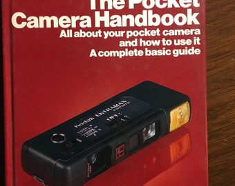Pode incluir: Um livro vermelho intitulado "The Pocket Camera Handbook" com uma câmera Kodak Ektramax preta e laranja na capa. O livro é um guia básico completo para usar uma câmera de bolso.