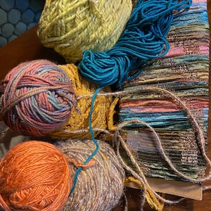 Könnte beinhalten: Eine Sammlung von Garn und Stoff in verschiedenen Farben, darunter Türkis, Orange, Braun, Gelb und Rosa. Das Garn liegt in Knäueln und Strängen vor, während der Stoff ein gestreiftes Band ist.