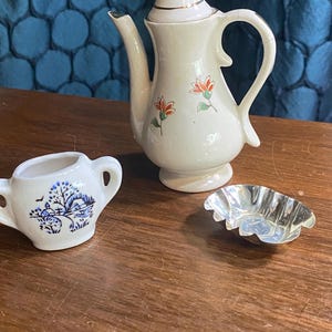 Może przedstawiać: Mały, kremowy ceramiczny imbryk z pozłacaną pokrywką i uchwytem, ozdobiony pomarańczowymi i zielonymi wzorami kwiatowymi. Obecna jest również pasująca mleczarka z niebiesko-białym krajobrazem i mała srebrna miseczka.