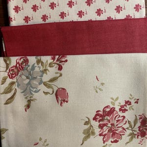 May include: Three fabric swatches. The top swatch is off-white with a repeating pattern of small red floral designs. The middle swatch is solid red. The bottom swatch is off-white with a floral pattern of red and blue flowers and green leaves.