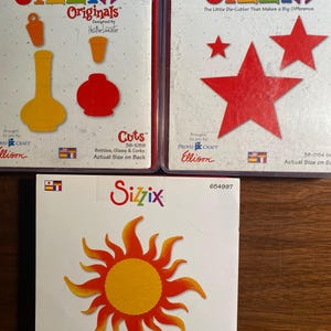 Może przedstawiać: Trzy zestawy do wycinania Sizzix do rękodzieła. Pierwszy zestaw zawiera czerwony, żółty i pomarańczowy wazon, czerwoną butelkę i czerwony słoik. Drugi zestaw zawiera trzy czerwone gwiazdy. Trzeci zestaw zawiera żółte słońce z pomarańczowymi promieniami. Zestawy są zapakowane w przezroczyste plastikowe pudełka z białym tłem i czarnym tekstem.
