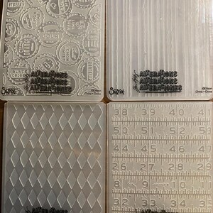 Puede incluir: Cuatro carpetas de relieve de plástico transparente con varios diseños. Una presenta tapas de botellas de refrescos, otra líneas verticales, una tercera un patrón de diamantes y la última una regla con medidas en centímetros. Cada carpeta tiene el texto "ALTERATIONS" y "Sizzix".