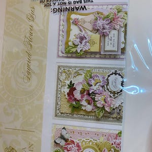 Może przedstawiać: Opakowanie wykrojnik&oacute;w warstwowych kwiat&oacute;w Anna Griffin. Opakowanie zawiera trzy ozdobne kartki z warstwowymi papierowymi kwiatami w odcieniach r&oacute;żu, fioletu i bieli. Kartki mają złote akcenty i tekst "Happy Birthday" i "For my friend, on this special day."