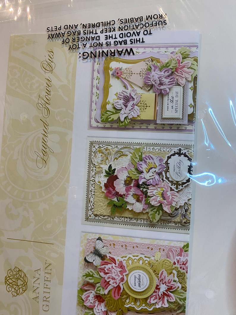 Może przedstawiać: Opakowanie wykrojnik&oacute;w warstwowych kwiat&oacute;w Anna Griffin. Opakowanie zawiera trzy ozdobne kartki z warstwowymi papierowymi kwiatami w odcieniach r&oacute;żu, fioletu i bieli. Kartki mają złote akcenty i tekst "Happy Birthday" i "For my friend, on this special day."