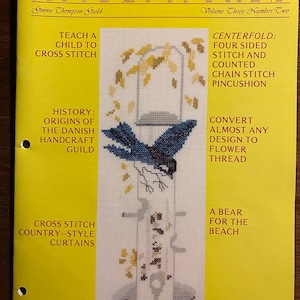 Puede incluir: Una portada de revista para Needlewords, Volumen Tres Número Dos, con un diseño de punto de cruz de un pájaro azul en una pajarera. La portada de la revista también incluye texto sobre enseñar a un niño a hacer punto de cruz, la historia del gremio artesanal danés, convertir casi cualquier diseño en hilo de flor y cortinas de estilo rústico de punto de cruz.