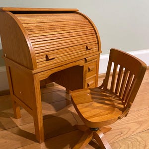 May include: A wooden roll-top desk and matching chair set. The desk features a curved, slatted top and small drawers. The chair has a slatted back and a swivel base. The set is made of light-colored wood.