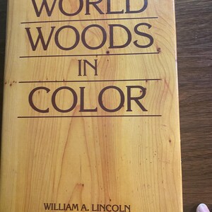 Op de afbeelding: Een boek getiteld "WORLD WOODS IN COLOR" door William A. Lincoln. De omslag heeft een houtnerfpatroon in tinten bruin en beige, met de titel in donkerbruine letters.