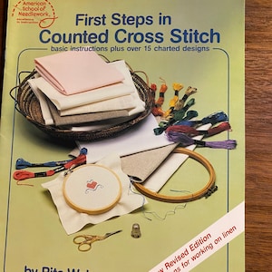 May include: A book titled "First Steps in Counted Cross Stitch" by Rita Weiss. The book cover features a basket of embroidery floss, a needlepoint hoop, and a close-up of a cross-stitch design. The book is a new revised edition with instructions for working on linen.
