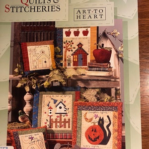 Puede incluir: Un libro que presenta una colección de colchas de calendario y patrones de bordado. La portada presenta una variedad de almohadas acolchadas coloridas con temas otoñales, incluyendo calabazas, manzanas y pájaros. El título "Calendar Quilts & Stitchery" y "Art-To-Heart" están impresos en la portada.