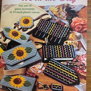 以下が含まれることがあります： 「It's in the Bag」というタイトルと「Five sets of purse accessories in 10-mesh plastic canvas」というサブタイトルが書かれた本の表紙。表紙には、ひまわり模様、キーホルダー、小さなハンドバッグなど、さまざまなカラフルなプラスチックキャンバスのプロジェクトが描かれています。
