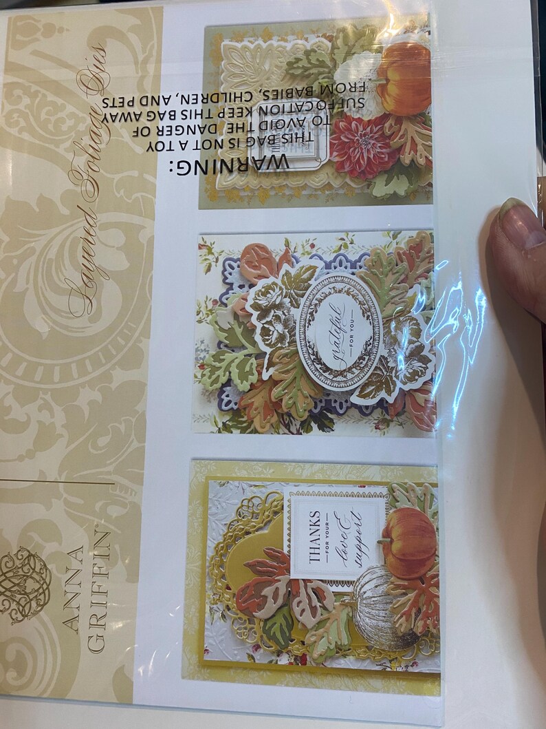 Może przedstawiać: Pakiet warstwowych wykrojnik&oacute;w do liści autorstwa Anny Griffin. Opakowanie zawiera trzy dekoracyjne projekty kart z elementami o tematyce jesiennej, w tym dynie, liście i akcenty kwiatowe. Karty zawierają słowa "grateful" i "thanks for your love & support."