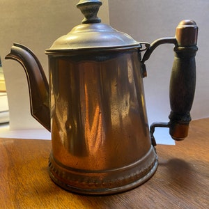 May include: A vintage copper teapot with a wooden handle and a decorative knob on the lid. The teapot has a worn, aged patina and a beaded trim around the base.