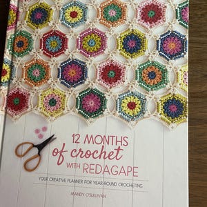 May include: A hardcover book titled 12 Months of Crochet with Redagape by Mandy O'Sullivan. The cover features a colorful pattern of crocheted hexagons in various hues joined by cream yarn, with an illustration of scissors and text.