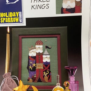 May include: A framed cross-stitch artwork showing three kings in red, purple, and gold robes and crowns. The text "WE THREE KINGS" is above the framed artwork. Gold stars, a crescent moon, and perfume bottles are in the foreground, with a lit candle.