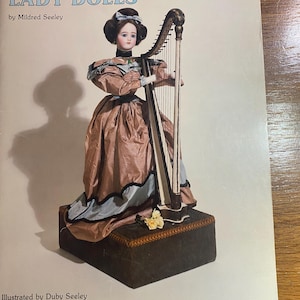 May include: The cover of "The Dollmaker's Workbook" features a lady doll playing a harp. The doll is dressed in a pink and blue gown and stands on a dark brown base. The title "LADY DOLLS" is printed in large blue letters.