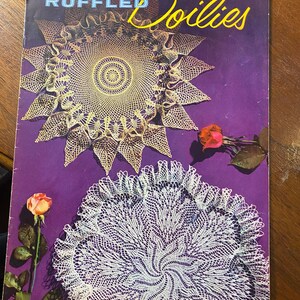 以下が含まれることがあります： 紫色の表紙に「RUFFLED DOILIES」という文字がスタイリッシュなフォントで書かれたヴィンテージの冊子。冊子のタイトルは「COATS & CLARK'S O.N.T. RUFFLED DOILIES BOOK No. 306 10¢」で、星形のデザインと渦巻き模様の2つのクロシェのドイリーが描かれている。