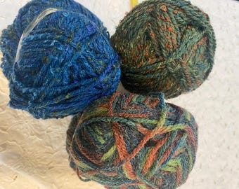 May include: Four balls of yarn in shades of blue, green, and orange. One ball is a bright blue with flecks of other colours. The other three balls are a mix of green, orange, and blue. The yarn has a textured, tweed-like appearance.