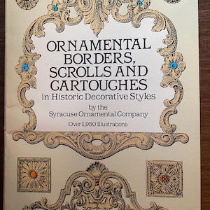 May include: A vintage book cover with the title "ORNAMENTAL BORDERS, SCROLLS AND CARTOUCHES" in black text. The cover features ornate gold-colored scrollwork and floral designs with blue and orange accents. The book is by the Syracuse Ornamental Company.