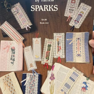May include: A collection of handmade cross-stitch bookmarks with lace trim and ribbon accents. Designs include floral, children's figures, and inspirational text. The book cover reads "STITCH-N-MARKS" by Patricia Sparks.