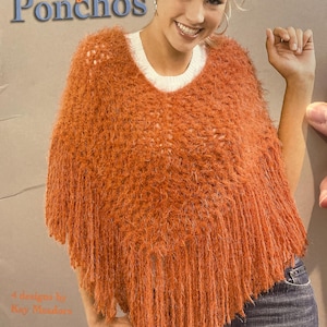 May include: An orange knitted poncho with a fringed hem, worn over a white sweater. The image features the text "Trendy Knit Ponchos" and "4 designs by Kay Meadors."