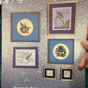 May include: A collection of framed cross-stitch designs with fantasy themes. The designs include a phoenix, a unicorn, a dragon, and a pegasus. The text "Figments of Fantasy" is at the top, with the designer's name at the bottom.