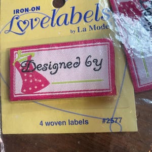 Może przedstawiać: Nanoszone żelazkiem tkane etykiety firmy La Mode. Etykiety są różowe z czerwoną obwódką i zawierają napis "Designed by" oraz grafikę sukienki. Opakowanie zawiera 4 etykiety i numer #2577.
