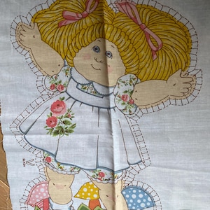 Può includere: Un pannello di tessuto con una bambola Cabbage Patch Kids. La bambola ha capelli gialli con fiocchi rosa, occhi blu e un incarnato rosato. L'abito è bianco con un motivo floreale e il disegno include funghi e fiori.