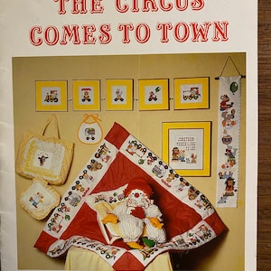 May include: A colorful cross-stitch pattern book titled "The Circus Comes to Town" by Pumpkin Patch Originals. The book features a variety of circus-themed designs, including a clown, a train, and a big top.