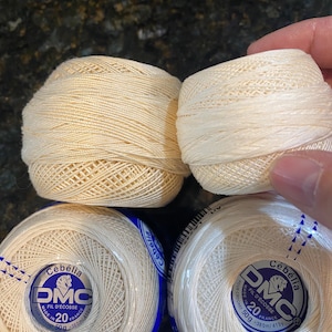以下が含まれることがあります： オフホワイトのDMCセベリアかぎ針編み糸の複数のスプール。スプールにはDMCのロゴと、糸が20番、50g、380m/415ヤードであることを示すテキストが記載されています。糸はボール状にしっかりと巻かれています。
