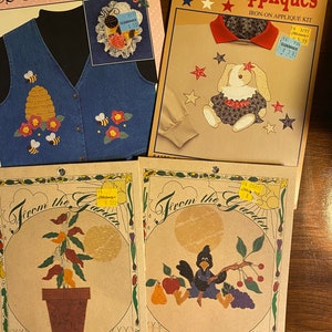 May include: Four different fabric applique kits. The first kit features a blue denim vest with a beehive and bees. The second kit features a tan shirt with a red collar and a white bunny with a red bow. The third kit features a chili pepper plant in a brown pot. The fourth kit features a crow sitting on a branch with fruit.