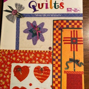Op de afbeelding: Een boek getiteld "Easy Quilts by Jupiter!" van Mary Beth Maison. De omslag toont een patchwork-quiltontwerp met een verscheidenheid aan kleuren en patronen, waaronder rood, geel en paars. Het boek maakt deel uit van een "Designer Series" en wordt uitgegeven door That Patchwork Place.