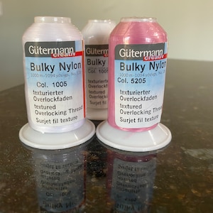 May include: Three spools of Gütermann Creativ Bulky Nylon thread. The spools are white, off-white, and pink. Each spool has the text "Gütermann Creativ Bulky Nylon" and the colour number. The white spool is colour 1005, the off-white spool is colour 100, and the pink spool is colour 5205.