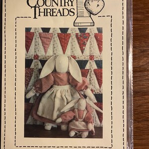 May include: A sewing pattern for making two country-style bunny dolls. The pattern is called "Timeworn Country Rabbits" and is number #110. The pattern features a larger bunny doll wearing a red and white gingham dress and a smaller bunny doll wearing a red and white gingham dress with a white apron.