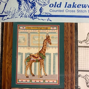 May include: A framed cross-stitch pattern of a giraffe on a carousel. The pattern is titled "Circa 1910 Dentzel Giraffe".