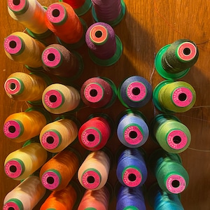 May include: A collection of 20 spools of thread in various colors, including red, orange, yellow, green, blue, purple, and pink. Each spool is labeled with the brand name "ISACORD" and the thread number. The spools are arranged in a circular pattern on a wooden surface.