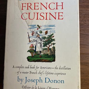 May include: A vintage cookbook titled "THE CLASSIC FRENCH CUISINE" with red and blue text. The cover features an illustration of a chef juggling food items. The author is Joseph Donon, Officier de la Légion d'Honneur.