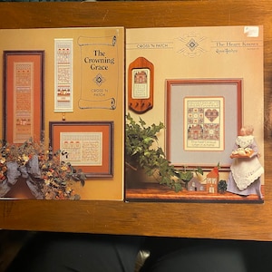Puede incluir: Dos patrones de punto de cruz, uno titulado "The Crowning Grace" y el otro titulado "The Heart Knows". Ambos patrones están enmarcados y presentan un tema de hogar.